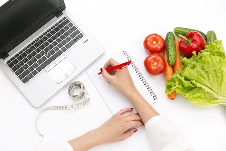 Person Writing Notes with a Laptop, Vegetables, and Measuring Tape
