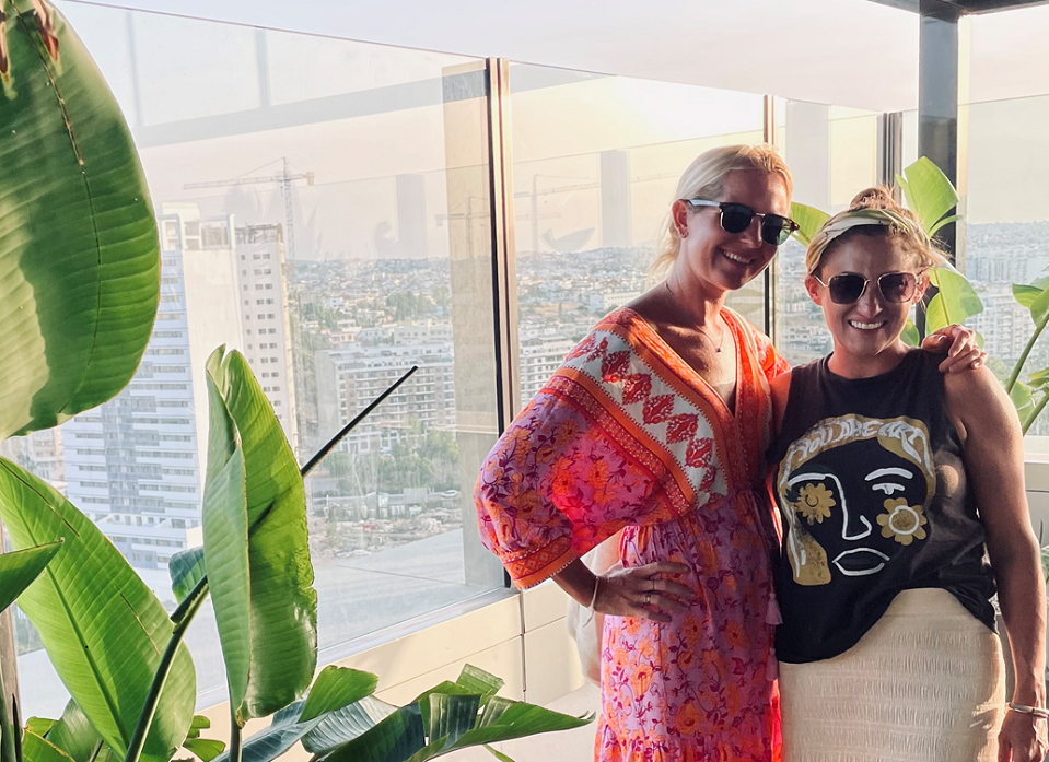 PJ's Coffee franchisee Aubry Miller and her sister in law posing next to each other in front of a window with a view of a city. A leafy plant frames them in the left corner.